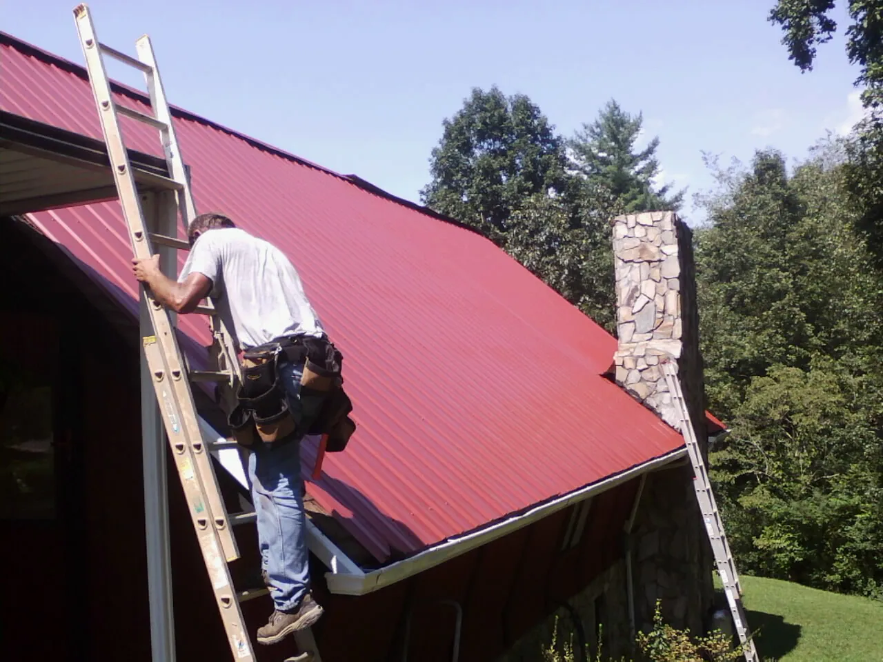 Roofer on a ladder beside a red metal roof and stone chimney — Soffit & Fascia Repair by Roofing Brothers in Amenia